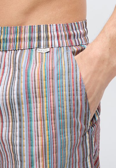 Χέρι άντρα στην τσέπη πολύχρωμων σορτς με κάθετες ρίγες, ελαστική μέση και μικρό υφασμάτινο ταμπελάκι μάρκας.