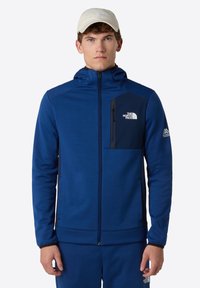 Blauwe zip-up jas met een capuchon en een borstzak in donkerblauw. Heeft een klein logo aan de linkerkant en elastische manchetten.