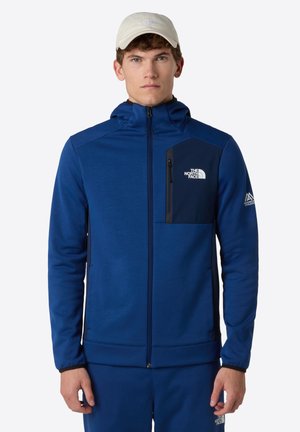 Blauwe zip-up jas met een capuchon en een borstzak in donkerblauw. Heeft een klein logo aan de linkerkant en elastische manchetten.