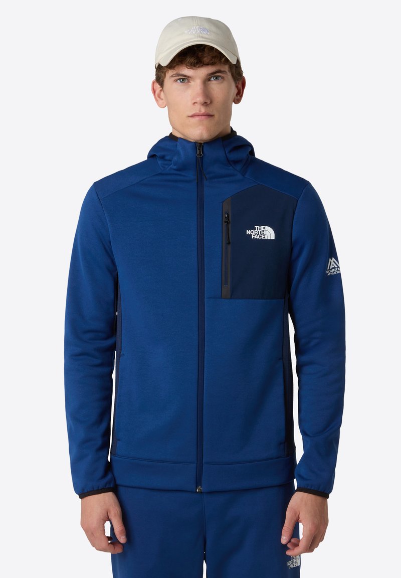 Blauwe zip-up jas met een capuchon en een borstzak in donkerblauw. Heeft een klein logo aan de linkerkant en elastische manchetten.