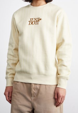 Personne portant un sweat crème à fines rayures avec un logo Nike marron et un pantalon beige, montrée du cou aux hanches.