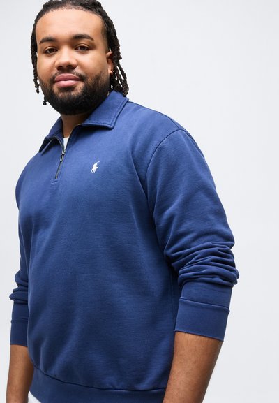 Άνδρας με πλεξουδάκια στα μαλλιά, φορόντας μπλε μπλούζα με μακριά μανίκια και φερμουάρ στη λαιμόκοψη, με ένα μικρό λευκό κεντητό λογότυπο στο στήθος.