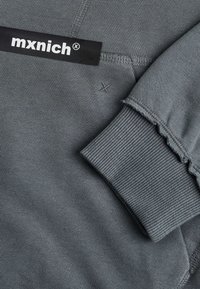 Siva dukserica s rebrastim manžetama i poderanim rubovima. Ima crnu etiketu s brandom "mxnich" na vrhu. Mekana, teksturirana tkanina.