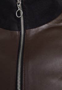 Brun læderjakke med en sølvlynlås og en ribstrikket sort krave. Glat tekstur med synlige syningsdetaljer langs sømmene.