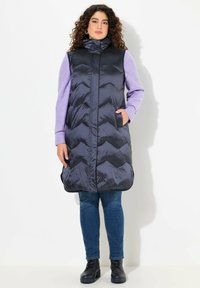 Lång marinblå pufferväst med hög krage, chevronmönster och sidofickor. Buren över en lila långärmad tröja och kombinerad med jeans och stövlar.