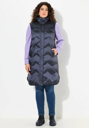Gilet matelassé long bleu marine avec col haut, motif en chevrons et poches latérales. Porté sur un pull long à manches longues lilas et associé à un jean et des bottes.