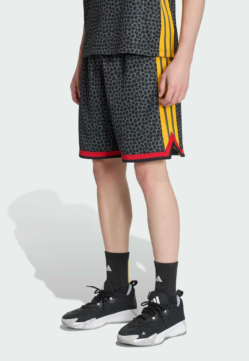 Personne portant un short noir à motifs avec des rayures rouges et jaunes, des chaussettes noires et des baskets noires Adidas debout sur un sol gris clair.