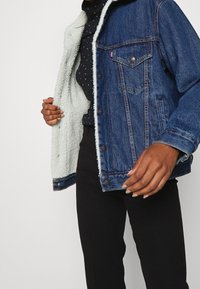 Džínová bunda s tmavě modrým exteriérem, bílou sherpa podšívkou, zapínáním na knoflíky a kapsami na hrudi. Pod ní je vidět černé oblečení.