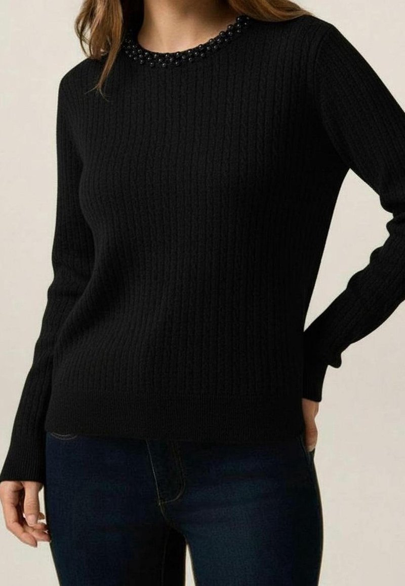 Femme portant un pull noir à manches longues côtelé avec un col orné de perles, associé à un jean bleu foncé, debout avec une main sur la hanche.