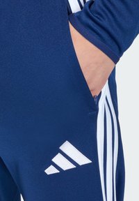 Αθλητική φόρμα σε ναυτικό μπλε χρώμα με φερμουάρ στην πλευρική τσέπη, με λευκές τρεις ρίγες και εμφανές λευκό λογότυπο adidas.