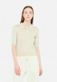 Polo maglietta beige a maniche corte con colletto, caratterizzata da quattro bottoni e un tessuto leggermente strutturato, abbinata a pantaloni bianchi.