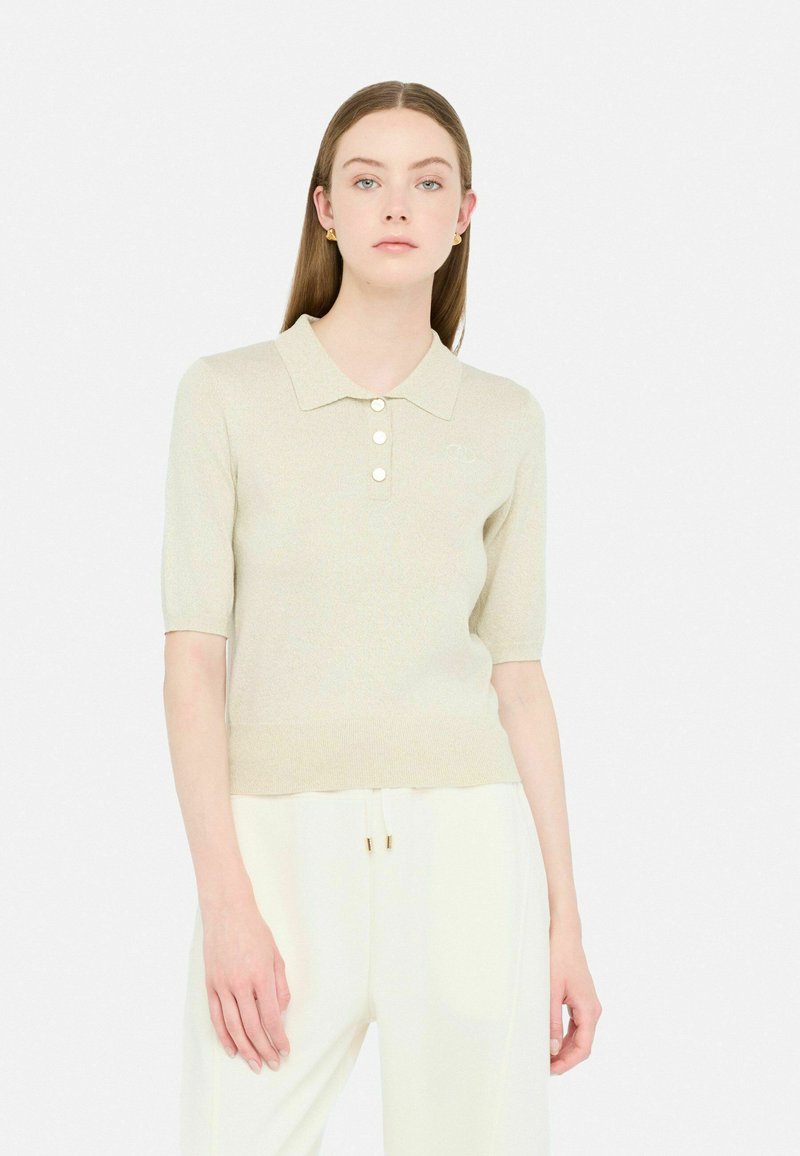 Polo maglietta beige a maniche corte con colletto, caratterizzata da quattro bottoni e un tessuto leggermente strutturato, abbinata a pantaloni bianchi.