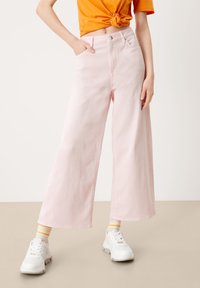 Jean rose à jambes larges avec taille haute, en coton, quatre poches, et fermeture par bouton ; porté avec des baskets blanches et des chaussettes rayées.