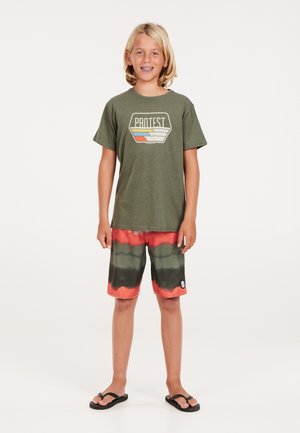 Grünes Baumwoll-T-Shirt mit einem grafischen "PROTEST"-Logo, kombiniert mit rot-grünen Batik-Badehosen und schwarzen Flip-Flops.