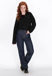 Zwarte trui met gesprenkeld patroon, oversized pasvorm en knopen op de schouders. Gepaard met rechte spijkerbroek in blauwe denim en zwarte schoenen.
