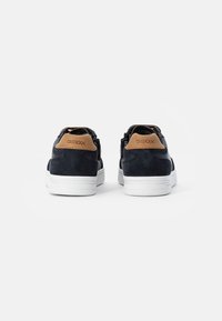 Zapatillas de ante negro con suelas de goma blanca, con un acento de piel marrón en el talón y detalles perforados en los laterales.