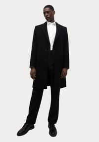 Abrigo de lana negro con botones grandes, combinado con una camisa blanca de cuello y pantalones negros. Unos zapatos de vestir negros brillantes completan el conjunto.
