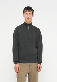 Cardigan grigio scuro con zip e colletto alto, realizzato in un tessuto morbido. Presenta maniche lunghe e polsini a costine, abbinato a pantaloni beige chiaro.