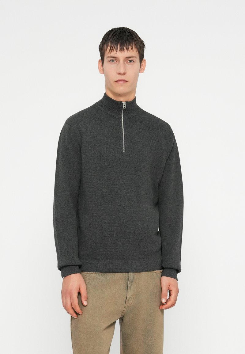 Cardigan grigio scuro con zip e colletto alto, realizzato in un tessuto morbido. Presenta maniche lunghe e polsini a costine, abbinato a pantaloni beige chiaro.