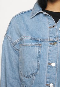 Ljusblå denimjacka med en framficka, silverknappar, bärs över en svart topp, visad från axel till midja.