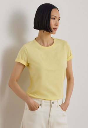 Rövid fekete hajú nő világos sárga rövid ujjú ingben és fehér nadrágban, zsebébe téve a kezét, jobbra nézve áll.