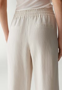 Lichtbeige broeken met een losse pasvorm, elastische tailleband en twee achterzakken met eenvoudige stikseldetails. Zacht, gestructureerd materiaal.