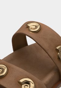 Καφέ λουράκι σανδάλια από σουέντ με χρυσά καρφάκια σε σχήμα κοχυλιού, κοντινό σε πάτο και πάνω λουράκι σε απλό φόντο.