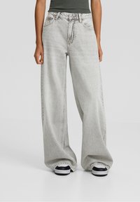 Personne portant un jean large gris clair et des baskets noir et blanc, les mains détendues le long du corps sur un fond uni.