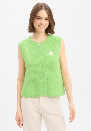 Jonge vrouw die een mouwloos limegroen gebreid spencer met knopen en lichtbeige broek draagt, glimlachend, staand tegen een witte achtergrond.