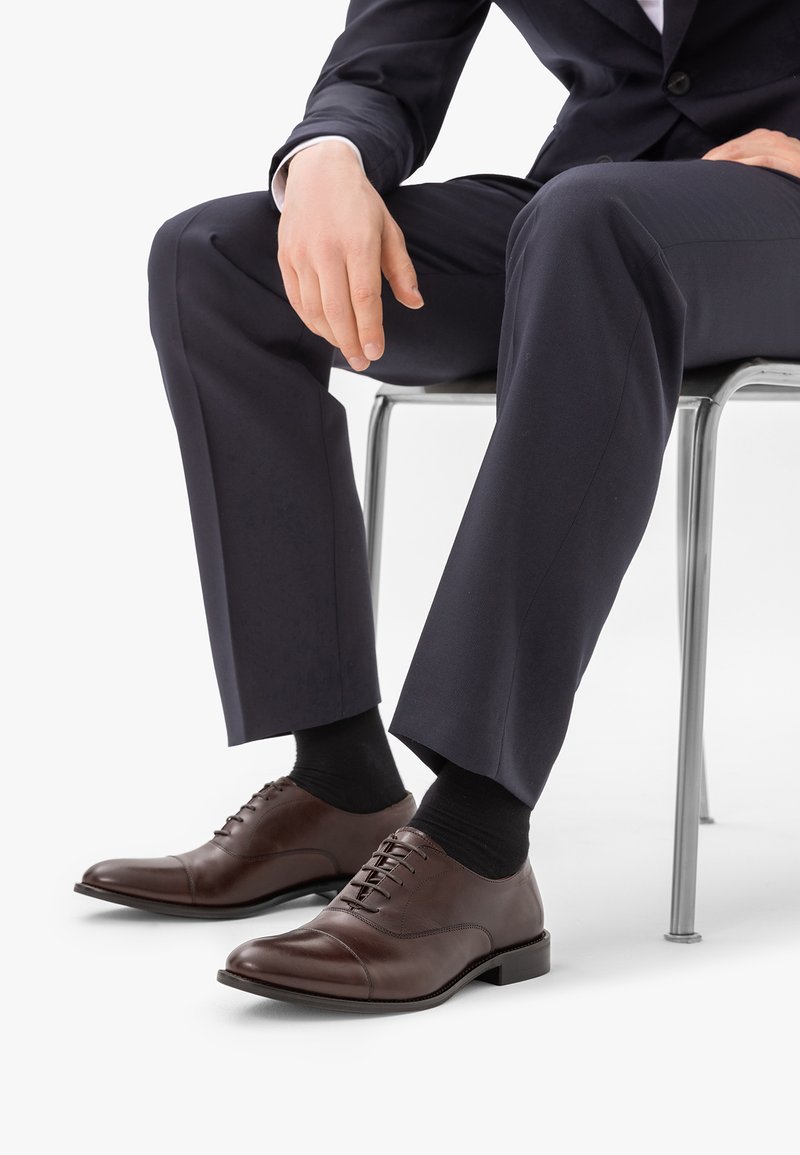 Homme portant un costume sombre et des chaussettes noires assis sur une chaise, montrant des chaussures habillées en cuir marron poli et une position des mains détendue.