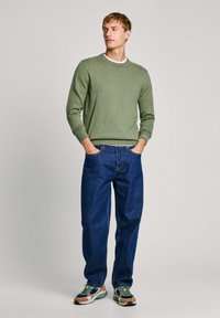 Suéter verde de punto con cuello redondo, combinado con jeans de mezclilla azul sueltos y zapatillas deportivas multicolores. Ajuste casual y diseño minimalista.