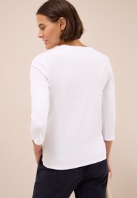 Camisa blanca de manga larga con cuello redondo y textura suave, combinada con pantalones oscuros. Se ve la parte trasera de la camisa.