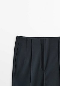 Zwarte high-waisted broek met plooien aan de voorkant en zijzakken, weergegeven tegen een witte achtergrond.