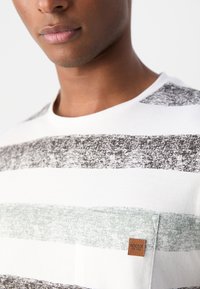 Camiseta blanca con rayas horizontales en gris, gris oscuro y verde menta, con un bolsillo en el pecho izquierdo y una etiqueta de marca marrón.