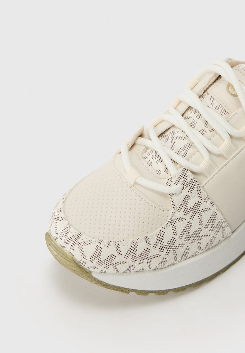 Chaussure de sport beige avec dessus en mesh comportant un motif. Embout doux et texturé, lacets blancs et semelle de couleur claire.