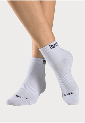 Pair of crossed legs wearing white ankle socks with "Bench" on cuffs and "sport" on toes against a plain light background.