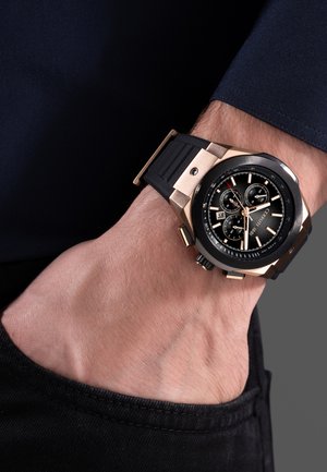 Muñeca de un hombre con un reloj cronógrafo negro y dorado con correa de goma negra, mano metida en el bolsillo de unos vaqueros negros.