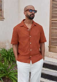 Short-sleeve, rust-colored shirt with embroidered sun patterns, white buttons, and a collared neckline, paired with light beige pants.