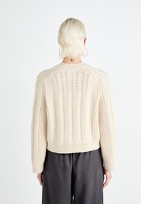Beige knitted sweater featuring a textured cable pattern, ribbed hem, and wide sleeves, displayed from the back against a plain background.