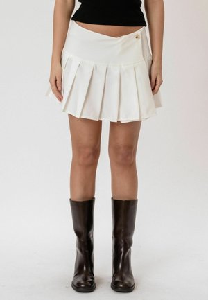 Mujer con una minifalda plisada blanca con detalle de botones y botas de cuero negras hasta la mitad de la pantorrilla, de pie contra un fondo liso.