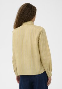 Femme avec des cheveux bouclés portant une chemise à manches longues jaune avec des rayures verticales blanches, vue de dos sur un fond uni.