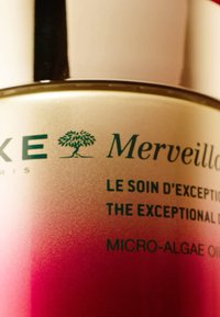 Bottiglia con gradiente rosa e dettagli in argento. Il testo include "Merveilleux", insieme al marchio e alle informazioni sul prodotto riguardanti l'olio di micro-alga.