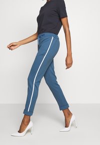 Kobieta w niebieskich zwężanych spodniach z białym paskiem po bokach i podwiniętymi mankietami, czarnej bluzce z krótkim rękawem oraz białych szpilkach, idąca.