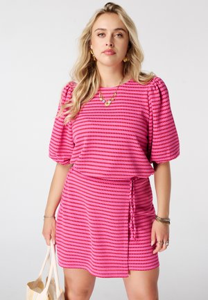 Blonde vrouw staat in een roze en rood gestreepte jurk met opgeblazen mouwen, gouden sieraden en houdt een beige schoudertas vast.