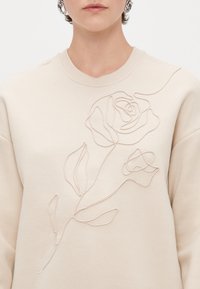 Μπεζ φούτερ με υφή, κεντημένο σχέδιο ρόζου με λεπτές γραμμές και στρογγυλή λαιμόκοψη. Απαλή, λείος ύφασμα.