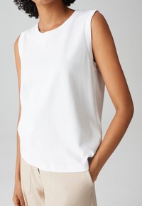 Weißes, ärmelloses Oberteil aus glattem Baumwollstoff mit rundem Halsausschnitt. Einfaches Design, leicht lockere Passform, mit Seitenschlitzen.
