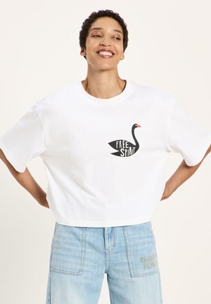 Person smiling wearing a white cropped t-shirt with a black swan and "Free Spirit" text, paired with light blue jeans with front pockets.