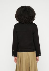 Črna jeans jakna z dolgimi rokavi, z zapenjanjem na sponke in gladkim teksturo. Ima prilagojen kroj in rahlo zaobljen zadnji rob.