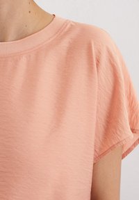 T-shirt de couleur pêcher avec un col rond, des manches courtes et un tissu légèrement texturé. L'ourlet et le col sont soigneusement finis.