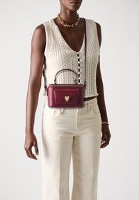 Dark-skinned person wearing cream cable-knit sleeveless top and pants, with gold chain necklace, bracelet, and small burgundy crossbody bag.